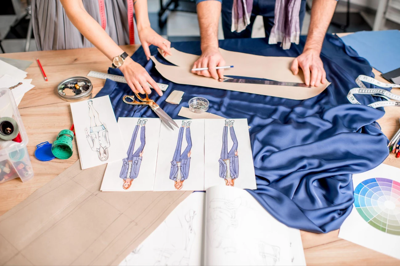 Fashion designers working over blue fabric with patterns and sketches on a worktable.
